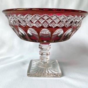May include: A pedestal bowl crafted from clear and deep red glass. The bowl features a wide, flared rim with a diamond-cut pattern above a row of circular indentations. It stands on a clear, multi-tiered stem and a square, ray-cut base.