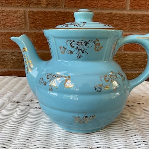 Puede incluir: Una tetera vintage de cerámica azul claro con tapa, asa y boquilla. La tetera está decorada con motivos florales dorados y ribetes. La tetera está sobre una superficie de mimbre blanca.