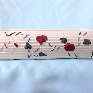 Puede incluir: Jardinera de cerámica color crema con textura acanalada y diseño floral pintado. La jardinera presenta detalles florales rojos y azul oscuro y líneas verticales negras en los extremos. Una pieza decorativa.