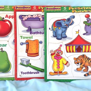 Op de afbeelding: Drie kleurrijke voorschoolse puzzels. Eén puzzel toont een rode appel en groene peer. Een andere toont een bad, handdoek en tandenborstel. De derde puzzel toont een circusscène met een olifant, clown en tijger.