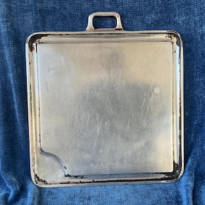 Peut inclure: Un plateau rectangulaire en métal vintage avec une poignée. Le plateau a une surface argentée avec des rayures et de l'usure, et un bord foncé. La poignée est petite et rectangulaire. Le plateau est probablement utilisé pour servir ou transporter des articles.