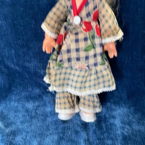 May include: A vintage doll wearing a plaid dress with red floral accents and a white collar. The doll's outfit includes matching plaid pants and white shoes. The doll has blonde hair and is set against a blue background.