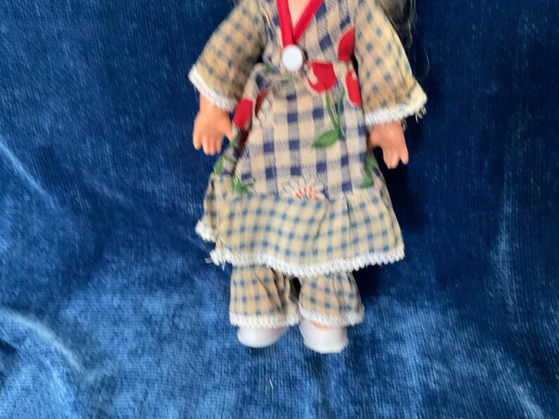 May include: A vintage doll wearing a plaid dress with red floral accents and a white collar. The doll's outfit includes matching plaid pants and white shoes. The doll has blonde hair and is set against a blue background.