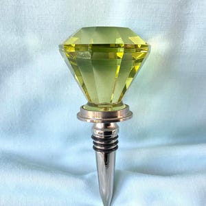 May include: A decorative bottle stopper with a faceted, diamond-shaped top in a pale yellow-green hue. The stopper has a silver-colored metal base and a tapered, stainless steel stem. The background is a light blue fabric.