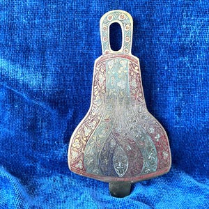 May include: An ornate, bell-shaped metal object with a handle. The object features intricate floral and geometric patterns in gold, red, and green against a silver background. The handle has a small opening. The object is resting on a blue surface.