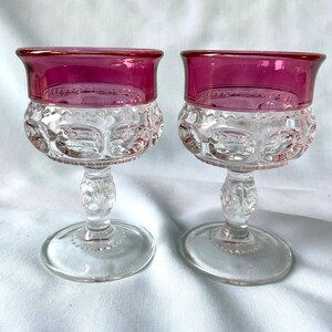 K&ouml;nnte beinhalten: Zwei antike Weingl&auml;ser aus klarem Glas mit rubinrotem Rand. Die Schale jedes Glases hat ein strukturiertes, gemustertes Design. Stiel und Fu&szlig; sind aus klarem Glas. Die Gl&auml;ser werden auf einer wei&szlig;en Oberfl&auml;che pr&auml;sentiert.