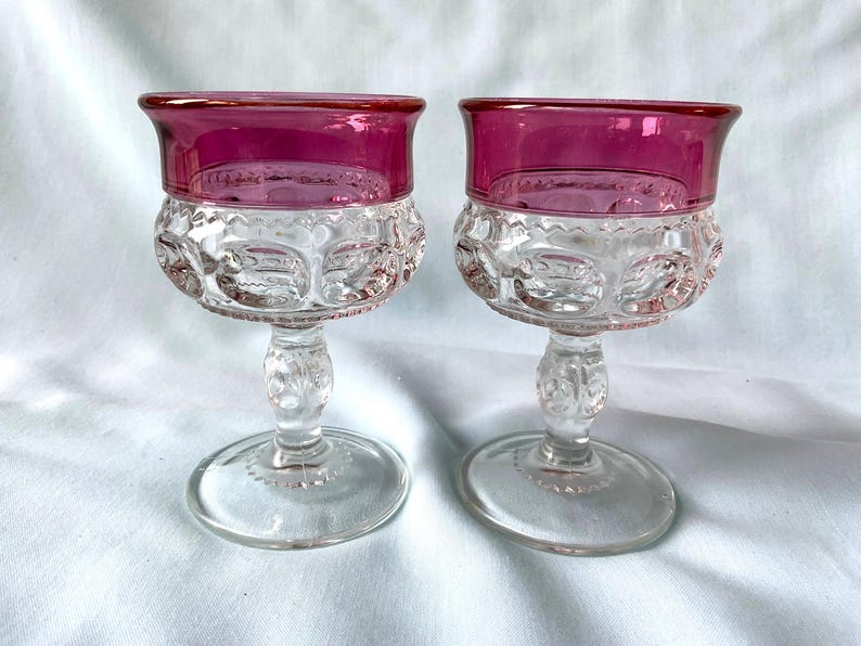 K&ouml;nnte beinhalten: Zwei antike Weingl&auml;ser aus klarem Glas mit rubinrotem Rand. Die Schale jedes Glases hat ein strukturiertes, gemustertes Design. Stiel und Fu&szlig; sind aus klarem Glas. Die Gl&auml;ser werden auf einer wei&szlig;en Oberfl&auml;che pr&auml;sentiert.
