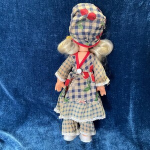 May include: A vintage doll wearing a checkered outfit with red cherry accents. The doll has blonde hair and is wearing a matching hat. The outfit includes a dress, pants, and a hat, all in a similar checkered pattern.