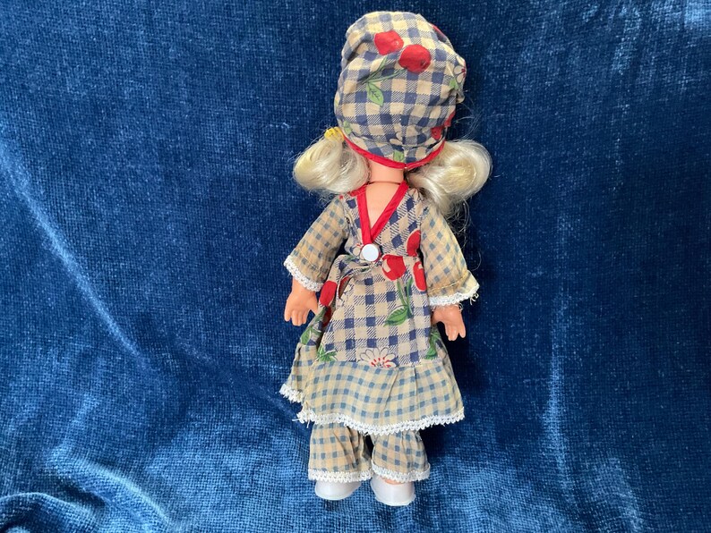 May include: A vintage doll wearing a checkered outfit with red cherry accents. The doll has blonde hair and is wearing a matching hat. The outfit includes a dress, pants, and a hat, all in a similar checkered pattern.