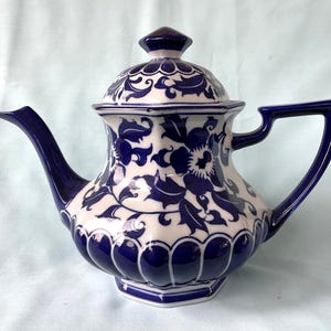 Op de afbeelding: Een keramische theepot met deksel, met een witte basis en ingewikkelde blauwe bloempatronen. De theepot heeft een gebogen tuit, een handvat en een geometrische basis. Het deksel is bekroond met een kleine, diamantvormige knop. Een decoratief keukenartikel.
