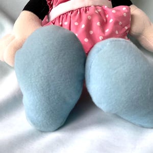 May include: A plush doll featuring light blue feet, a pink dress with white polka dots, and beige arms. The doll has black details and is set against a white background.