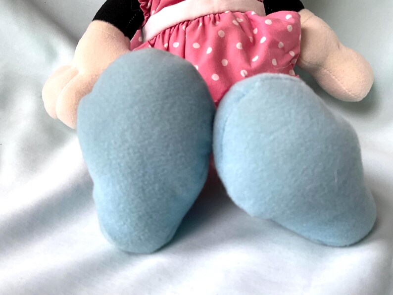 May include: A plush doll featuring light blue feet, a pink dress with white polka dots, and beige arms. The doll has black details and is set against a white background.