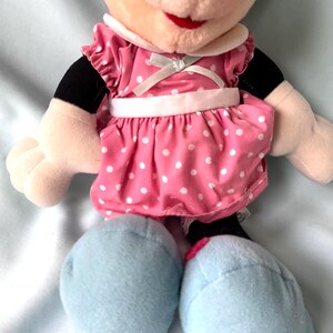 May include: A plush Minnie Mouse doll. The doll has a pink dress with white polka dots, a white collar, and light blue shoes. The doll's face has a red mouth and black eyes. The arms are beige with black sleeves.