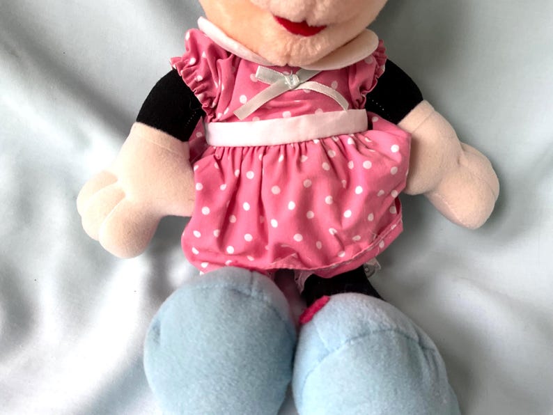 May include: A plush Minnie Mouse doll. The doll has a pink dress with white polka dots, a white collar, and light blue shoes. The doll's face has a red mouth and black eyes. The arms are beige with black sleeves.