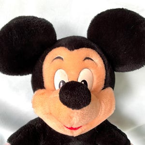 Puede incluir: Un peluche de Mickey Mouse con grandes orejas negras, una nariz negra y una cara color melocot&oacute;n. El juguete tiene una sonrisa roja y est&aacute; hecho de un material suave y difuso. Un personaje cl&aacute;sico de Disney.