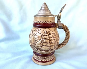May include: A ceramic beer stein featuring a relief of a sailing ship and coastal buildings. The stein has a metallic conical lid with a thumb lever, a rope-textured handle, and a dark red band at the base with the text Fine Bark Staysail.
