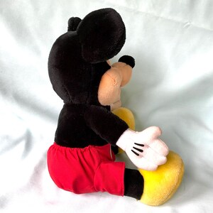 Puede incluir: Un peluche de Mickey Mouse, con cabeza y orejas negras, cuerpo negro, pantalones cortos rojos, zapatos amarillos y guantes blancos. El juguete est&aacute; en posici&oacute;n sentada, mostrando su dise&ntilde;o y colores cl&aacute;sicos.