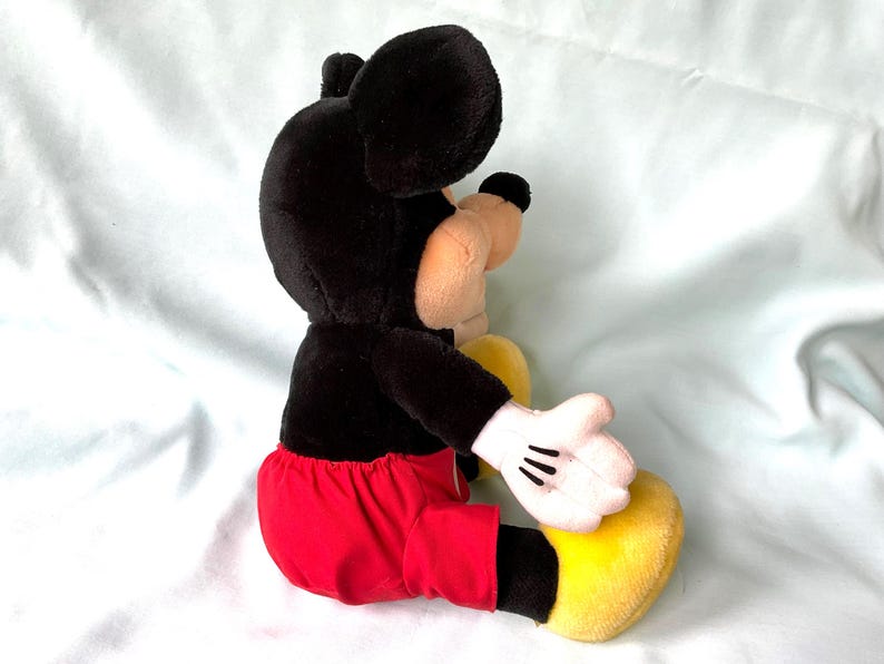 Puede incluir: Un peluche de Mickey Mouse, con cabeza y orejas negras, cuerpo negro, pantalones cortos rojos, zapatos amarillos y guantes blancos. El juguete est&aacute; en posici&oacute;n sentada, mostrando su dise&ntilde;o y colores cl&aacute;sicos.
