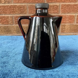 May include: A glossy, dark brown ceramic pitcher with a handle and a black lid. The pitcher has a unique, angular design and is set against a brick wall and a blue surface. The pitcher is approximately 8 inches tall.