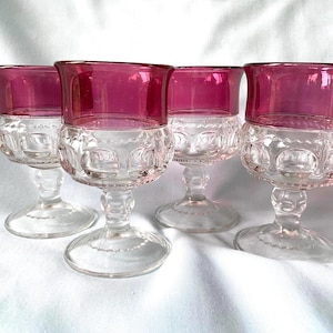May include: Four clear glass goblets with a decorative pattern on the lower half and a vibrant magenta rim. The goblets have a classic stem and base, set against a white background.