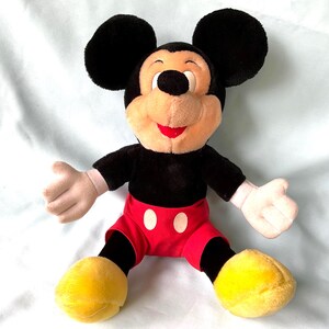 Puede incluir: Un mu&ntilde;eco de peluche de Mickey Mouse con grandes orejas negras, cuerpo negro, pantalones cortos rojos con botones blancos y zapatos amarillos. El mu&ntilde;eco tiene una cara beige, guantes blancos y una boca roja. El mu&ntilde;eco est&aacute; sentado con los brazos abiertos.