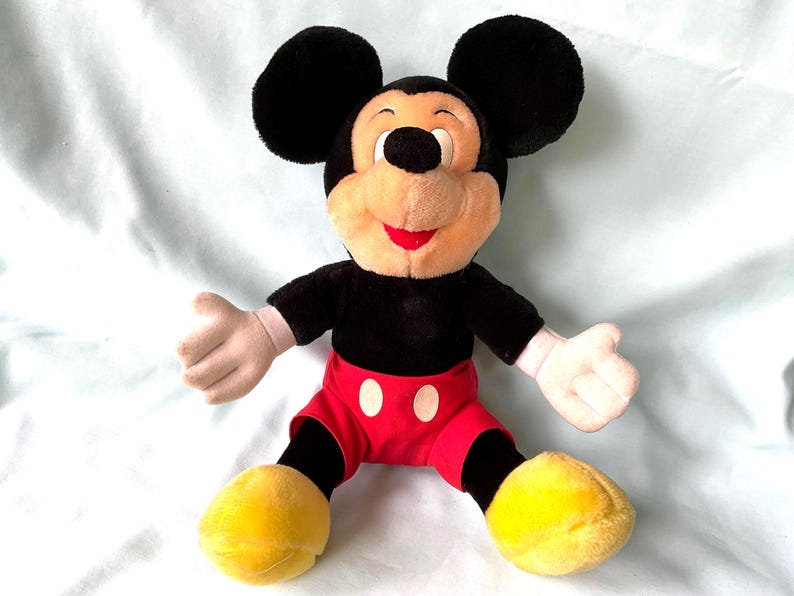 Puede incluir: Un mu&ntilde;eco de peluche de Mickey Mouse con grandes orejas negras, cuerpo negro, pantalones cortos rojos con botones blancos y zapatos amarillos. El mu&ntilde;eco tiene una cara beige, guantes blancos y una boca roja. El mu&ntilde;eco est&aacute; sentado con los brazos abiertos.