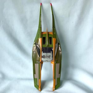 May include: A green and orange toy spaceship with two long, upward-pointing fins. The spaceship has a sleek, aerodynamic design with orange and gold accents. The tips of the fins are red. The toy appears to be made of plastic.