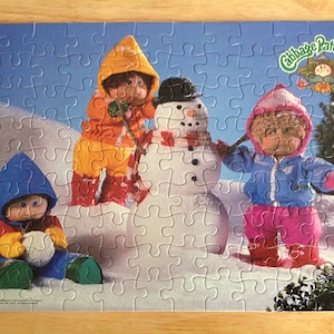 May include: A completed Cabbage Patch Kids jigsaw puzzle featuring four dolls building a snowman in a snowy landscape. The dolls wear colorful winter outfits, including red, blue, orange, and pink. The puzzle has a blue sky background.