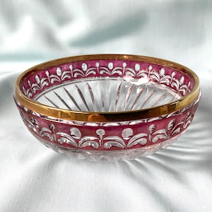 Op de afbeelding: Een ronde kristallen kom met een heldere glazen basis met verticale geslepen facetten. De bovenrand is versierd met een diepmagenta band, ingewikkelde witte bloemachtige geëtste patronen en een gepolijste goudkleurige metalen rand.