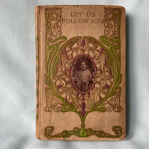 May include: Antique book titled "Let Us Follow Him" with an embossed floral design in green and purple on a tan cover. A portrait is centered within the design. The book's cover shows signs of age and wear.