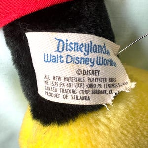 Puede incluir: Primer plano de un juguete de peluche vintage con gorra roja, pelaje negro y cuerpo amarillo. Una etiqueta dice "Disneyland Walt Disney World" y "&copy; Disney". La etiqueta tambi&eacute;n enumera los materiales y el fabricante.