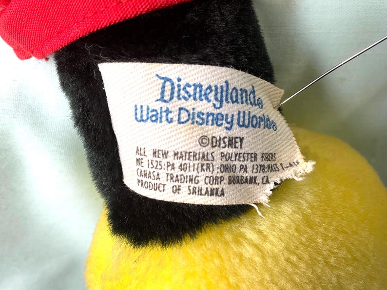 Puede incluir: Primer plano de un juguete de peluche vintage con gorra roja, pelaje negro y cuerpo amarillo. Una etiqueta dice "Disneyland Walt Disney World" y "&copy; Disney". La etiqueta tambi&eacute;n enumera los materiales y el fabricante.