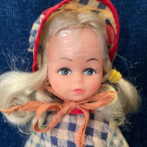 May include: A vintage doll with blonde hair styled in pigtails, blue eyes, and a red and checkered bonnet. The doll wears a checkered dress and a bow around its neck. A classic toy.