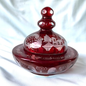 Könnte beinhalten: Eine tiefrote Glasschale mit passendem Deckel, verziert mit detaillierten Gravuren von Bäumen, Gebäuden und Wirbelmustern. Der Deckel hat einen gestuften, abgerundeten Knauf und der Boden ist mit einem wiederkehrenden Spiralmotiv versehen.