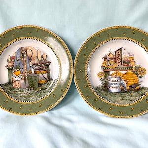 Puede incluir: Dos platos decorativos con borde verde y una detallada escena de jardín. Uno presenta herramientas de jardinería y un sombrero, el otro una casa para pájaros y una colmena. Los platos tienen un borde punteado amarillo y blanco.