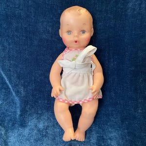 May include: A vintage baby doll with a surprised expression, wearing a white dress with pink gingham trim and a matching bib. The doll has blonde hair, blue eyes, and rosy cheeks. The doll is made of plastic.