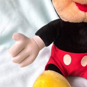 Puede incluir: Mu&ntilde;eco de peluche de Mickey Mouse con torso negro, pantalones cortos rojos con botones blancos y pies amarillos. El mu&ntilde;eco tiene cara y manos color canela y boca roja. El brazo del mu&ntilde;eco est&aacute; extendido.