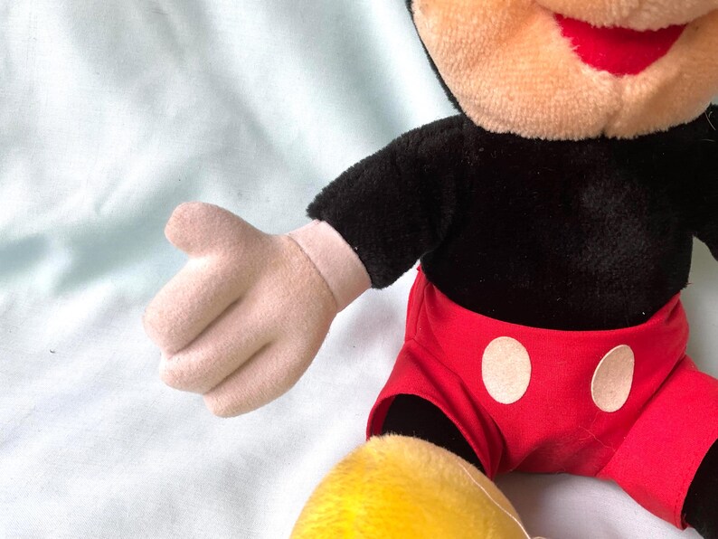 Puede incluir: Mu&ntilde;eco de peluche de Mickey Mouse con torso negro, pantalones cortos rojos con botones blancos y pies amarillos. El mu&ntilde;eco tiene cara y manos color canela y boca roja. El brazo del mu&ntilde;eco est&aacute; extendido.