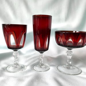 Peut inclure: Trois pièces de verrerie rouge foncé avec des pieds et des bases transparents. L'ensemble comprend un verre à vin, une flûte haute et une coupe large, présentant tous un motif arqué distinct à la base du calice où le verre rouge rencontre le pied.