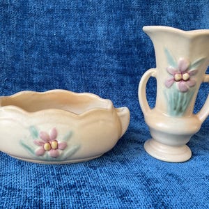Puede incluir: Dos jarrones de cerámica color crema. Uno es un cuenco pequeño y festoneado con asas, y el otro es un jarrón con la parte superior acampanada y dos asas. Ambos tienen un diseño floral rosa y verde.