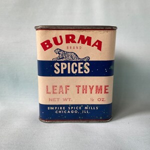 Może przedstawiać: Puszka przypraw vintage Burma Brand Leaf Thyme. Prostokątna puszka ma kremowe tło z czerwonym i niebieskim tekstem oraz logo tygrysa. Tekst brzmi "BURMA BRAND SPICES LEAF THYME WAGA NETTO 14g."