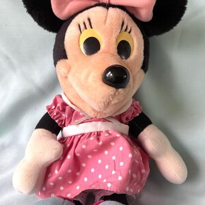 May include: A plush Minnie Mouse doll. The doll has a black head with large black ears and a pink bow. She wears a pink dress with white polka dots and a white belt. Her face is peach-colored with large yellow eyes and a black nose.