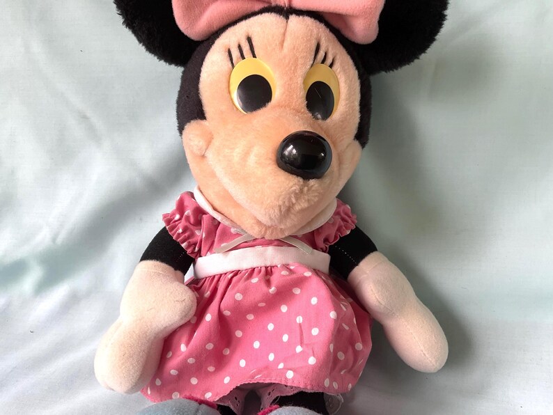 May include: A plush Minnie Mouse doll. The doll has a black head with large black ears and a pink bow. She wears a pink dress with white polka dots and a white belt. Her face is peach-colored with large yellow eyes and a black nose.