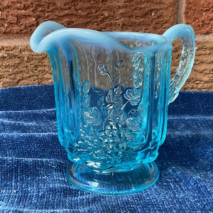 May include: A light blue opalescent glass creamer pitcher featuring a raised relief pattern of grapevines and leaves. The pitcher has a scalloped rim, a curved handle, and a circular base, showcasing a translucent finish with a milky white edge.