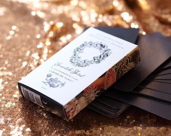 May include: A white box with black text and illustrations of sea life. The box reads "Seaside Tarot Le Tarot Enchanté de la Mer Cartes de Tarot". The box is sitting on top of a deck of black tarot cards.