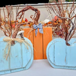 May include: Three decorative wooden pumpkins in a fall theme. Two are light blue with a distressed finish, and one is orange. Each has a twig stem and is adorned with raffia or a gingham bow. Fall decor.