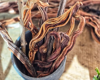 May include: A collection of decorative driftwood branches in a dark gray container. The branches display a variety of colors, including light brown, dark brown, and tan. The wood has a textured, layered appearance, with some branches exhibiting a twisted, curved shape.