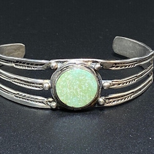May include: A silver cuff bracelet with a central, round, light green stone. The bracelet has three parallel bands with etched designs and small silver accents. The open ends allow for easy wear.