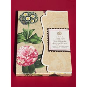 May include: A decorative box featuring floral illustrations. The box has a beige background with a floral design and a label that reads "ANNA GRIFFIN More Flower Pt Decoupage Die Cuts". The box is on a red surface.