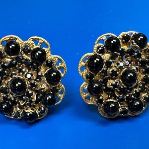 May include: A pair of vintage-style clip-on earrings. Each earring features a gold-toned filigree base, adorned with clusters of black, round, and faceted beads. The earrings have a floral design and are set against a blue background.